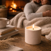 Lit nude concrete candle on a wooden table with a couple cuddled together by a cosy fire in the background