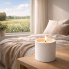 Lit white concrete candle on a bedside table with morning light and a wildflower field visible through the window
