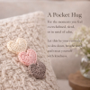 Three hand-crocheted heart pocket hugs resting on a soft knitted pillow with a calming message about slowing down and self-kindness.